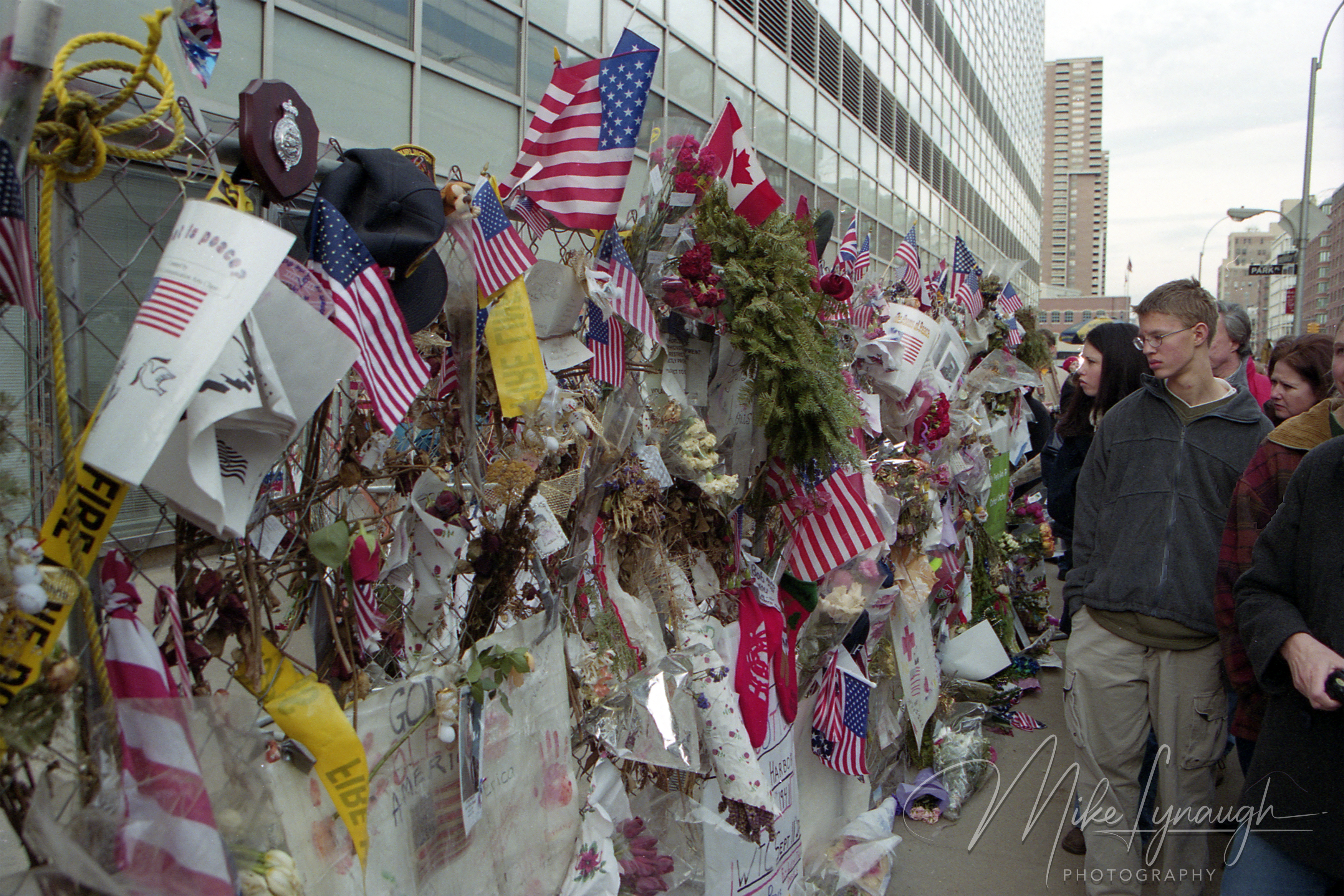 A chain-link fence filled with American flags, flowers, handwritten notes, and badges as visitors walk past near Ground Zero in late 2001.