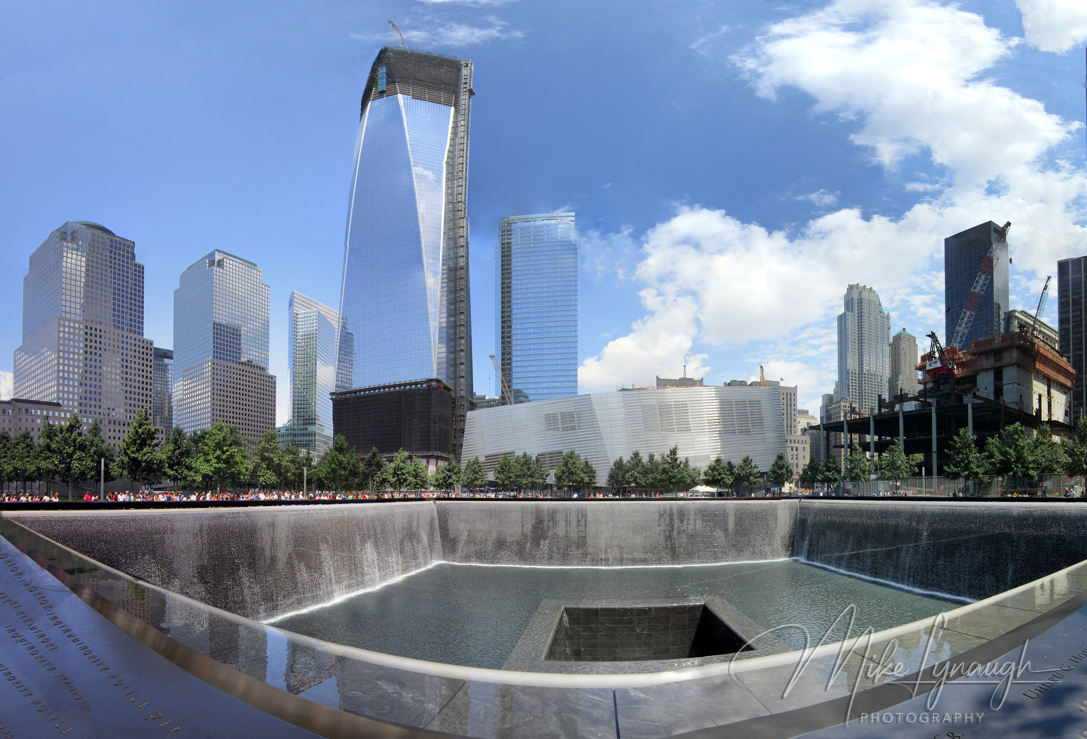 An ultra-wide fisheye panoramic view of the North 9/11 Memorial pool surrounded by visitors, swamp white oak trees, and the rising skyscrapers of the World Trade Center site.