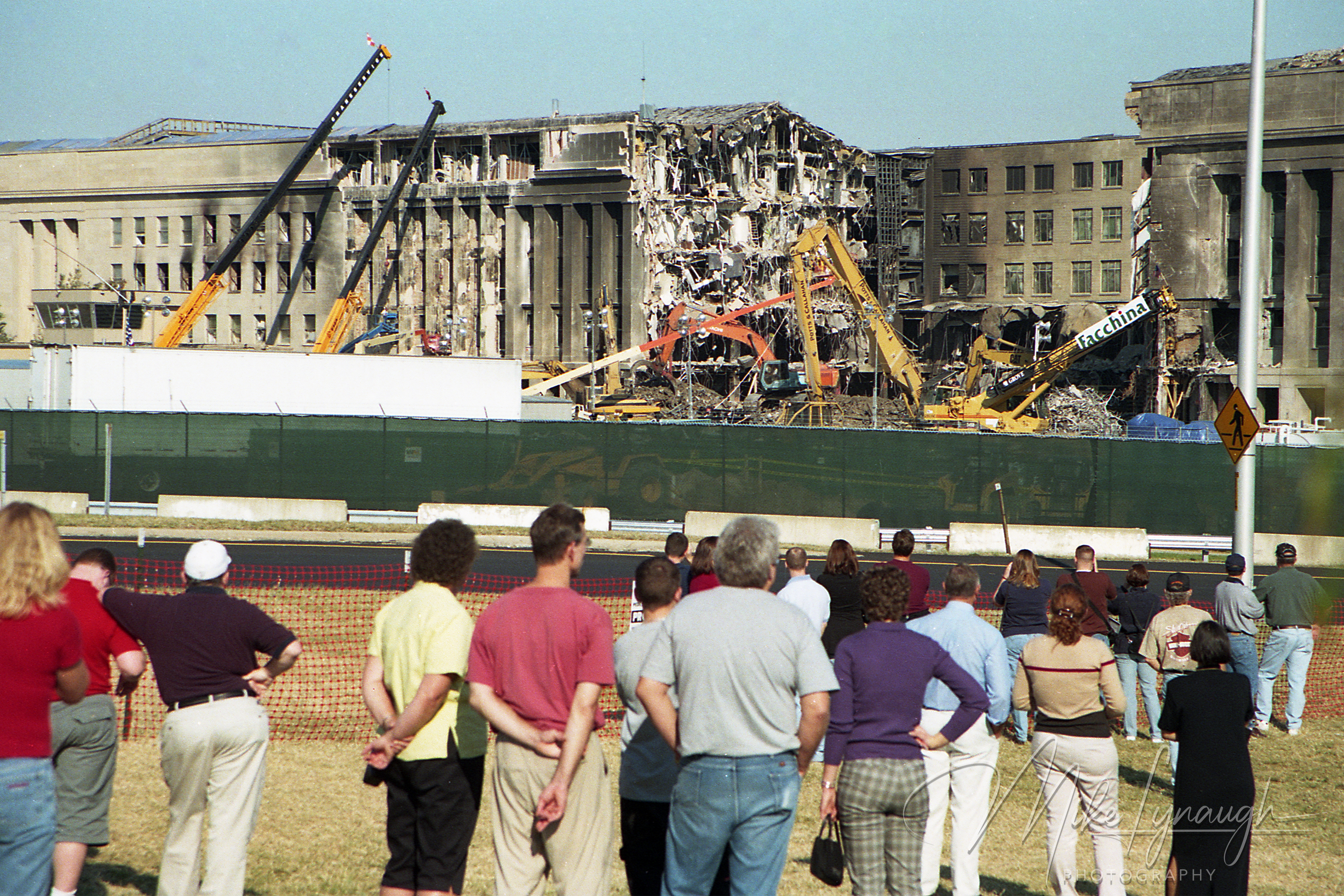 A crowd of citizens watching demolition cranes and heavy machinery clearing the damaged Pentagon structure from behind a perimeter fence