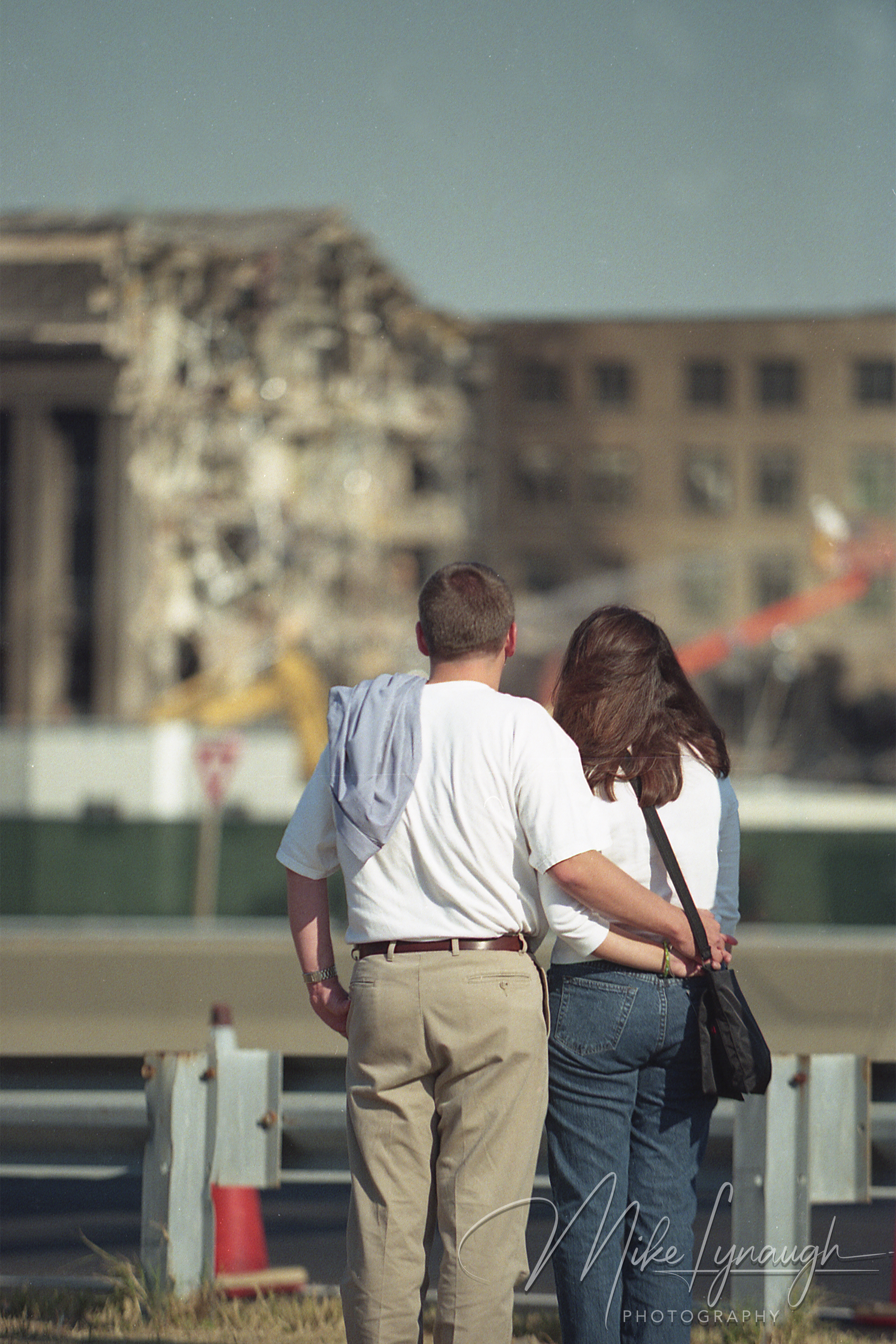 Rear view of a man and woman embracing as they look toward the softly blurred, damaged Pentagon facade in the background