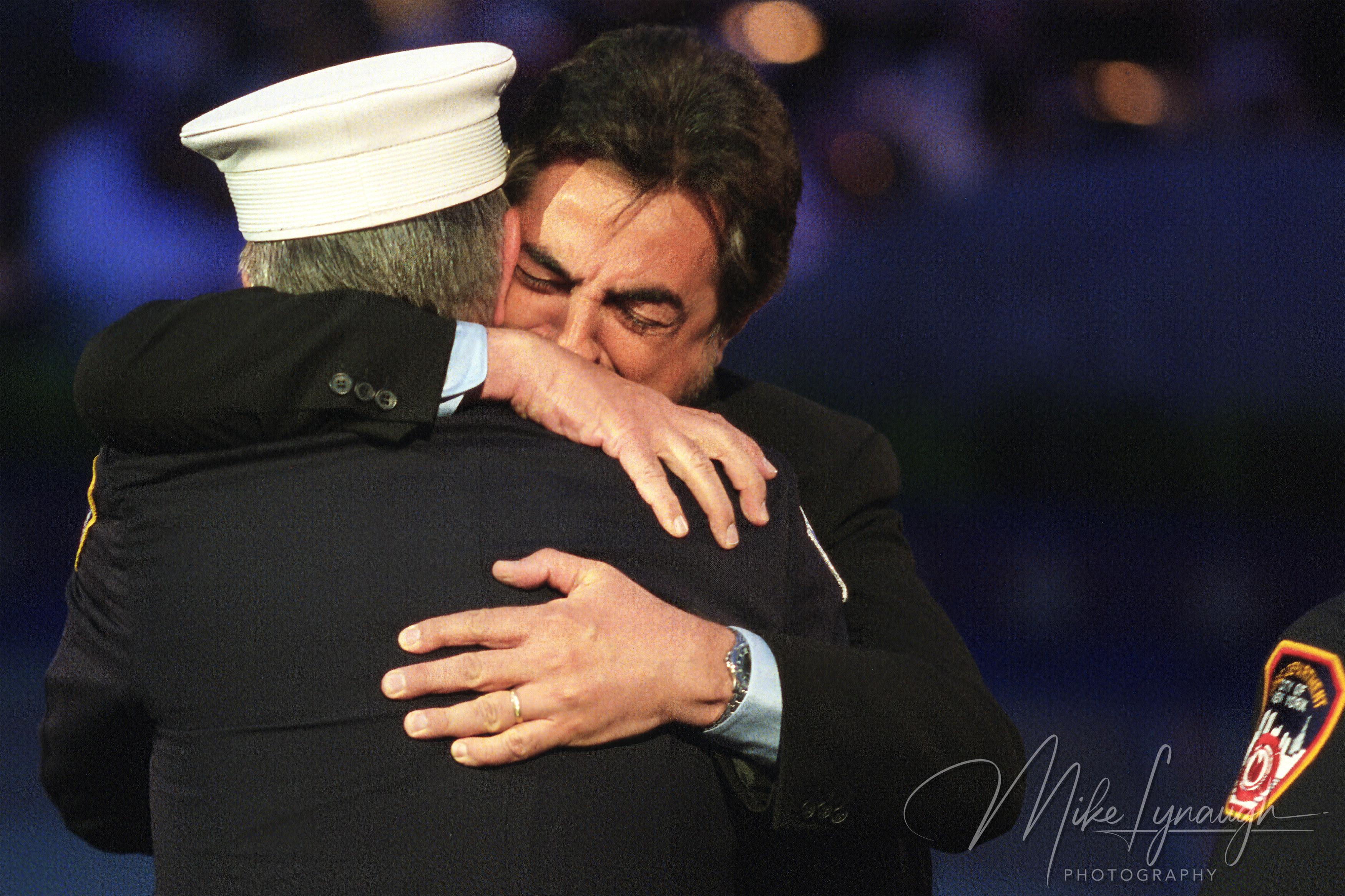 An emotional photograph of actor Joe Mantegna hugging retired FDNY Captain John Vigiano from behind on stage during the 2002 National Memorial Day Concert.