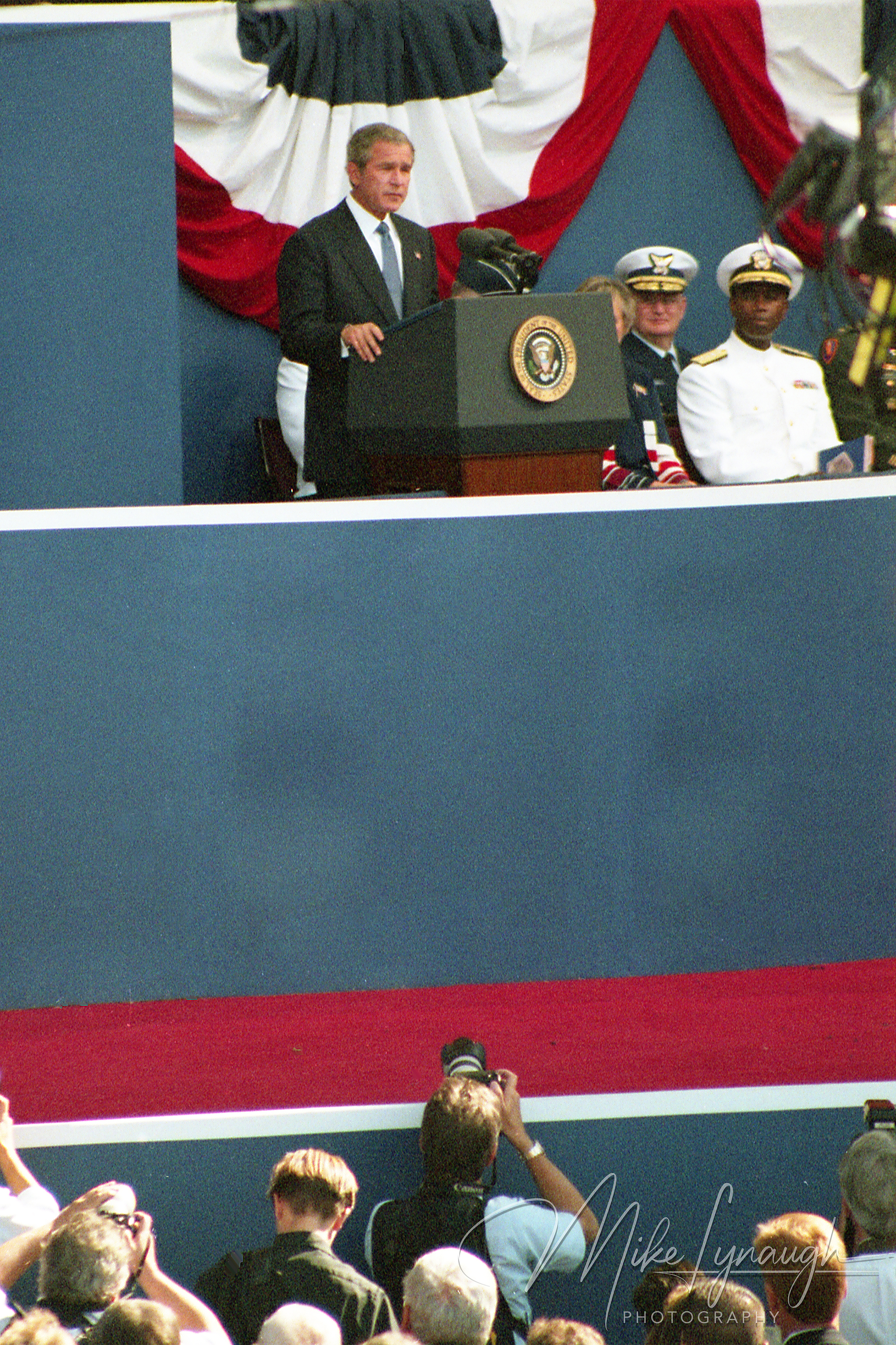 A vertical photo showing President George W. Bush speaking from a podium at an outdoor ceremony with red, white, and blue bunting in the background and photojournalists in the foreground