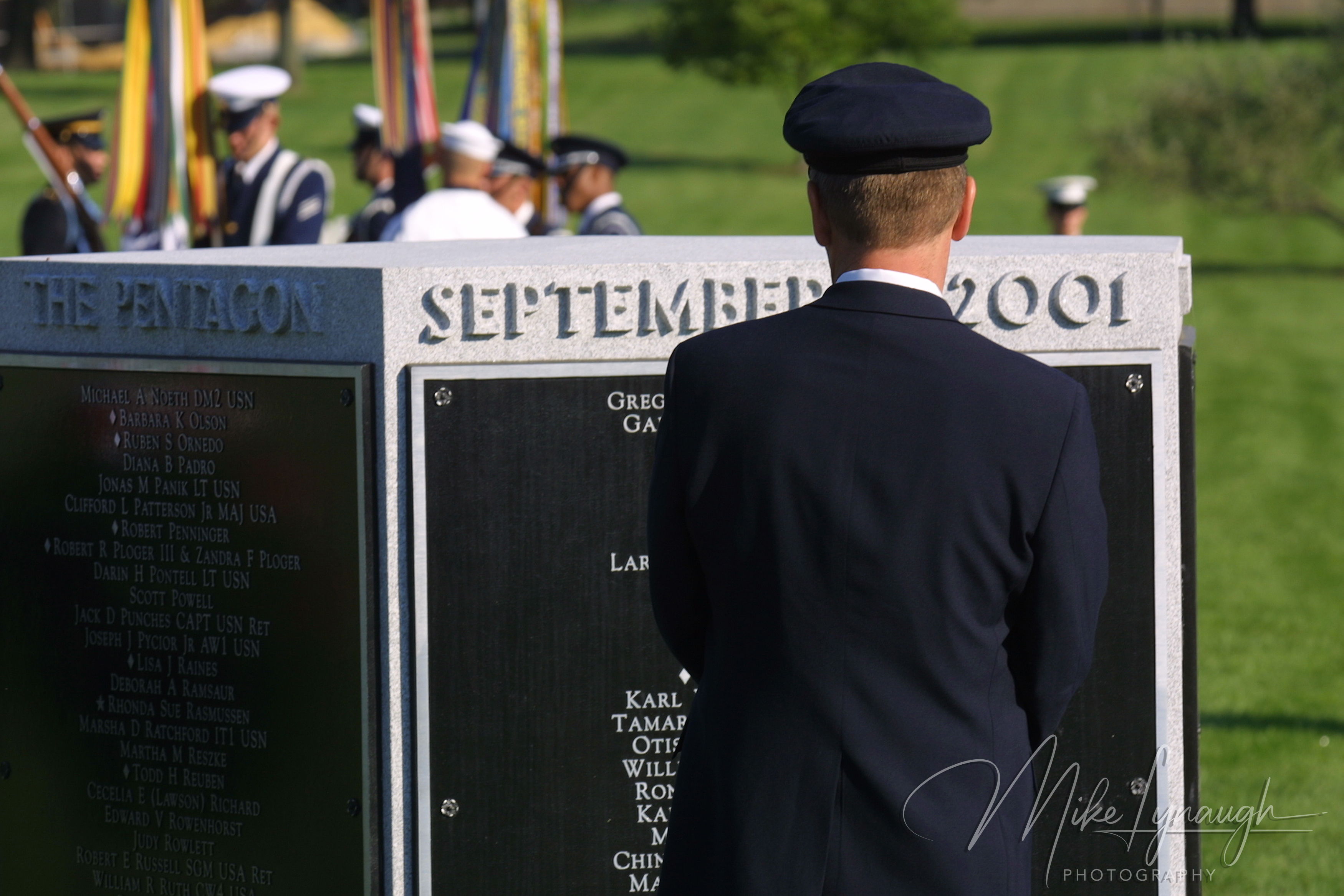Rear view of an American Airlines pilot in a dark uniform cap and blazer standing silently in front of the pentagonal 9/11 memorial marker at Arlington National Cemetery