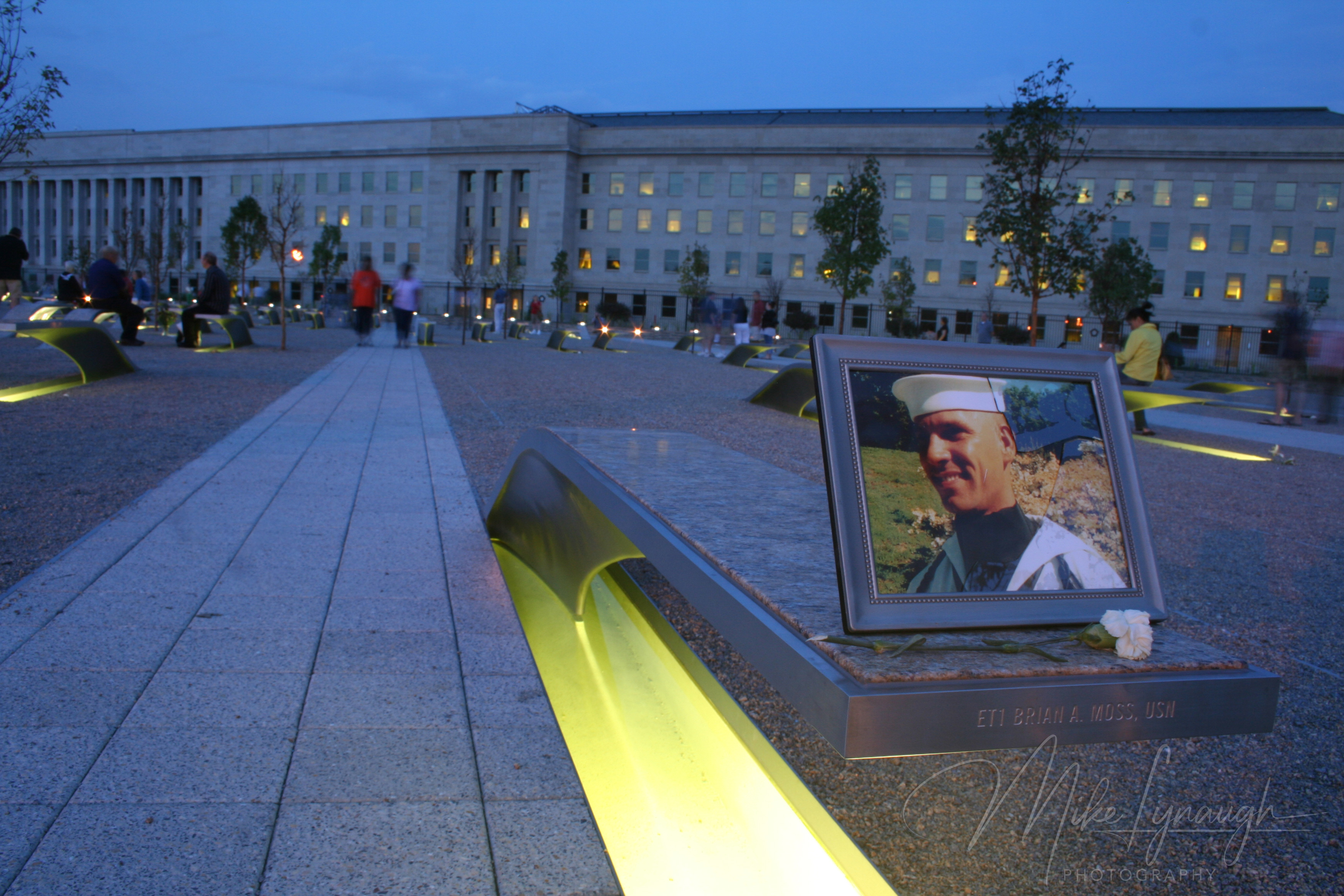 An illuminated memorial bench at dusk engraved with 'ET1 BRIAN A. MOSS, USN' holding a framed photo of a smiling sailor in uniform and a white rose, with the Pentagon building lit up in the background.