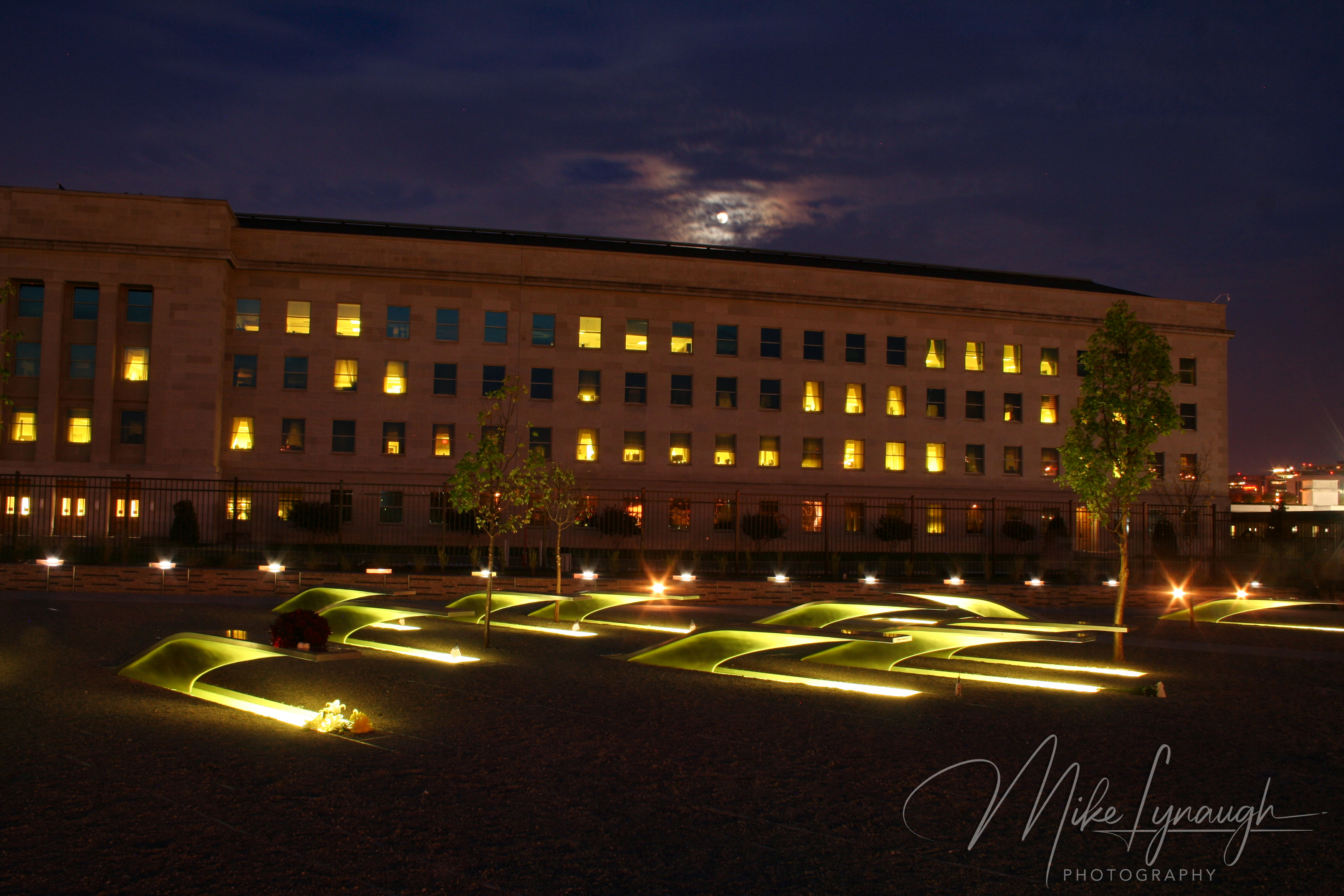 A night view of the National Pentagon Memorial showing glowing cantilevered benches over illuminated pools under a moonlit sky with the lit windows of the Pentagon building in the background.
