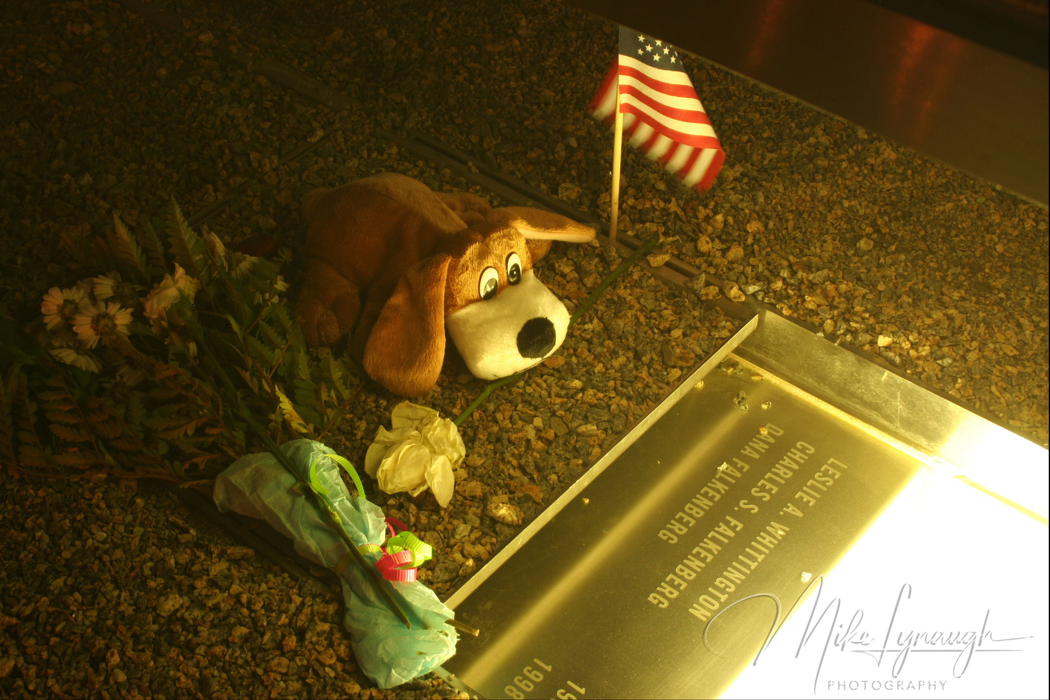 A close-up view of an illuminated memorial bench base engraved with the names Leslie A. Whittington, Charles S. Falkenberg, and Dana Falkenberg, with a plush stuffed dog, flowers, and an American flag placed beside it.