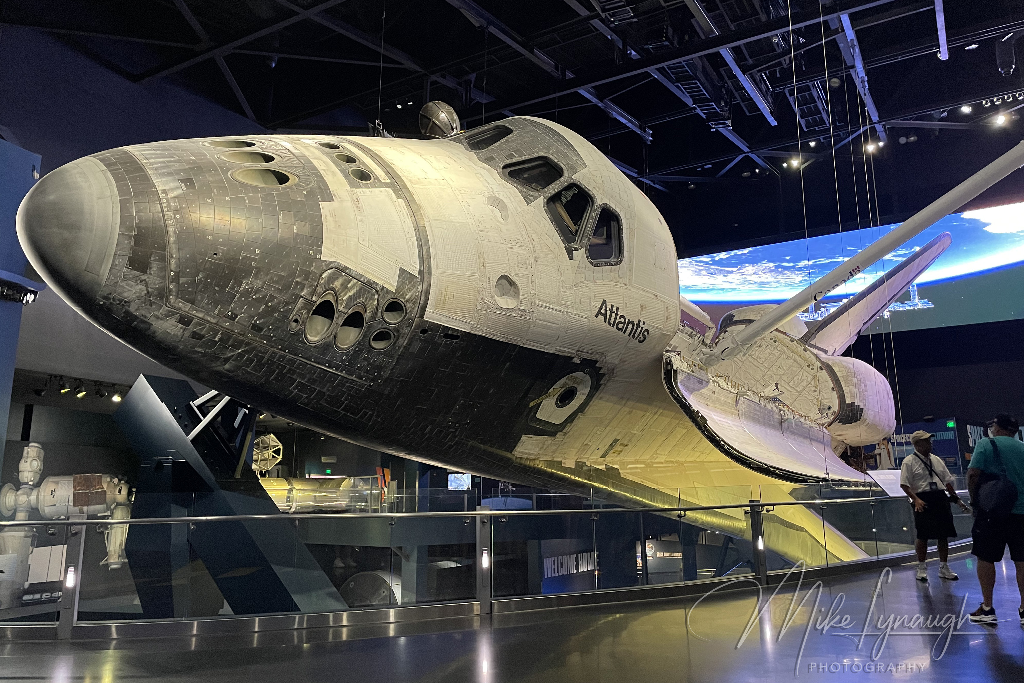 An interior museum photograph showing visitors admiring the authentic Space Shuttle Atlantis (OV-104) at the Kennedy Space Center Visitor Complex in Florida.