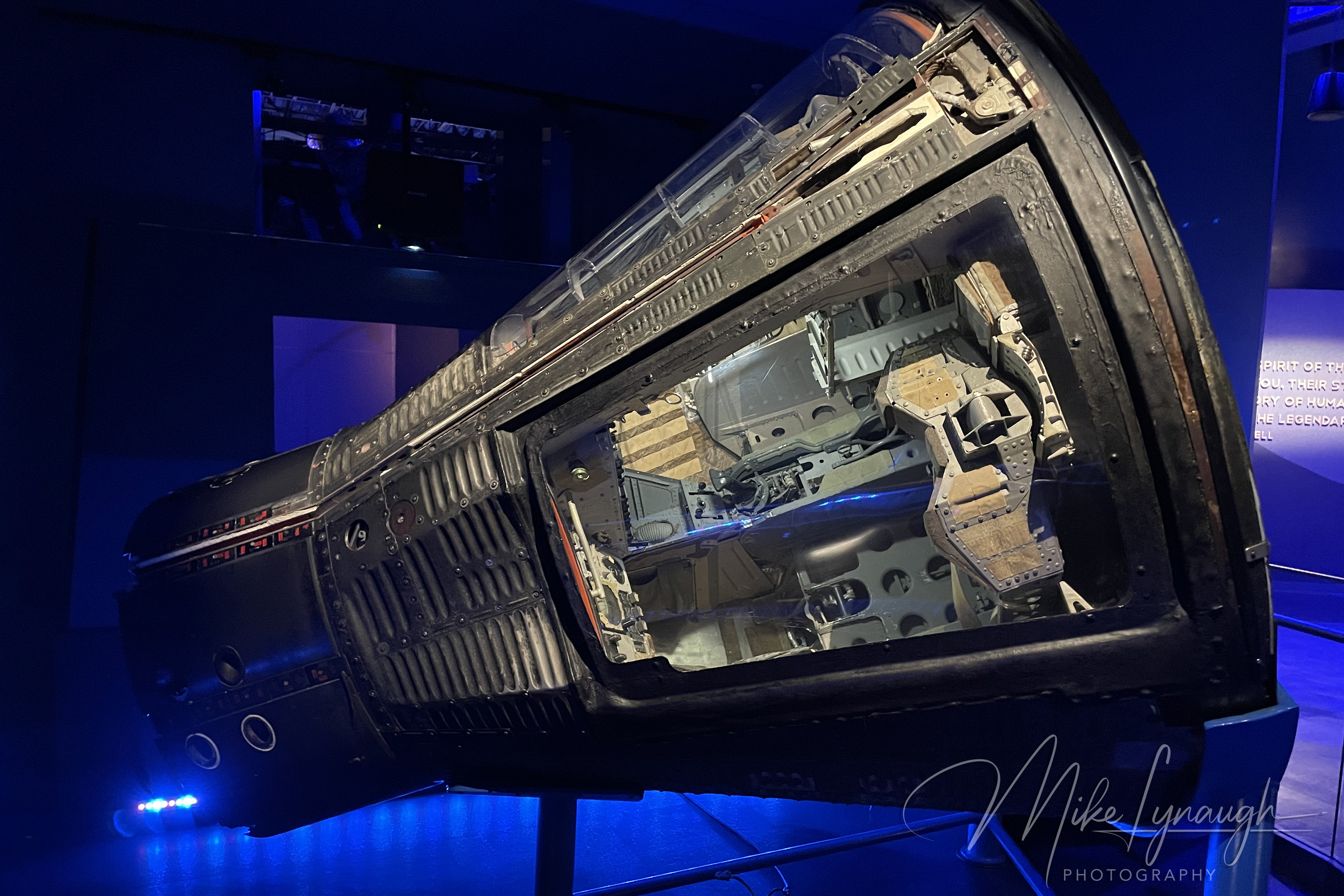 An interior museum photograph showing the authentic, flown Gemini 9A (Gemini IX-A) spacecraft capsule on display under blue atmospheric lighting inside the Heroes & Legends attraction at NASA's Kennedy Space Center.