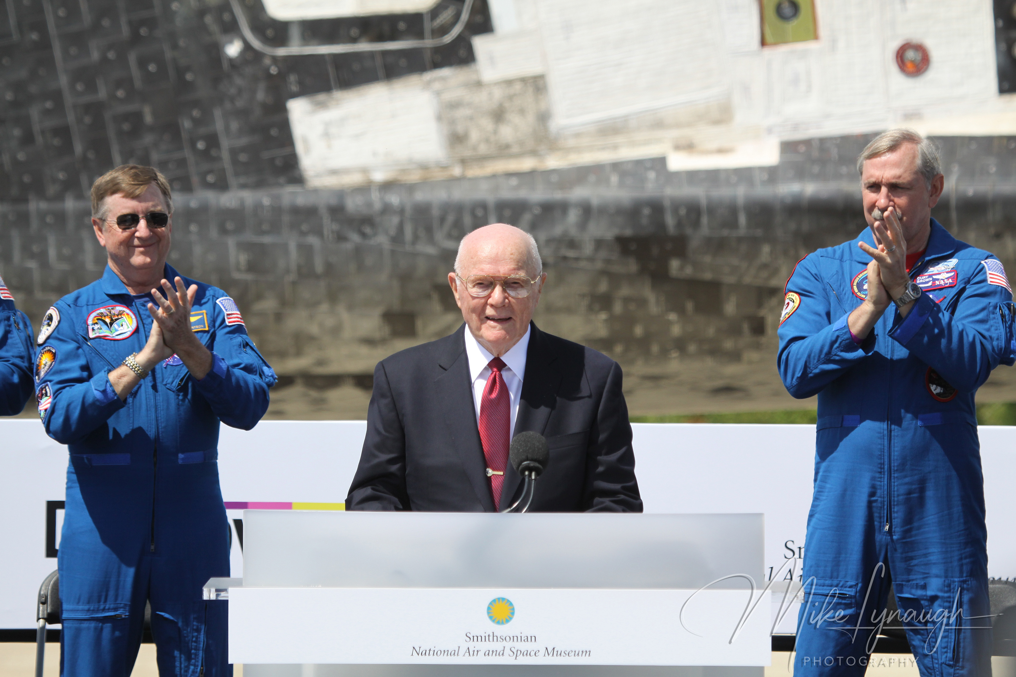 A medium portrait photograph capturing former astronaut and U.S. Senator John Glenn speaking at the Smithsonian podium at the Steven F.